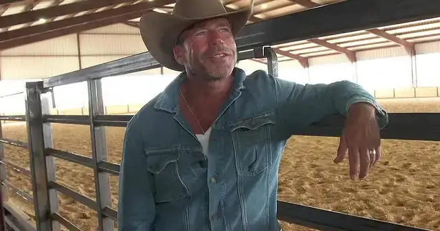 Screenwriter and director Taylor Sheridan in a denim jacket, white undershirt, and cowboy hat, leaning against a metal fence inside a large, covered equestrian riding arena.