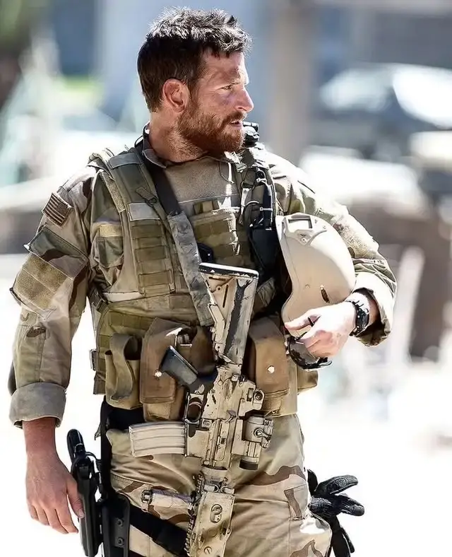 Actor Bradley Cooper, portraying Chris Kyle in American Sniper, wearing full desert camouflage tactical gear and holding a helmet in his right hand.