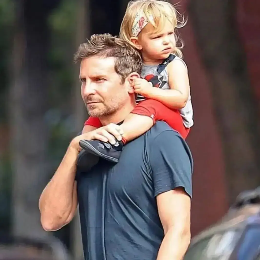 Actor Bradley Cooper carrying his daughter, Lea De Seine, on his shoulders as she holds onto his ear and looks away, both dressed casually outdoors.