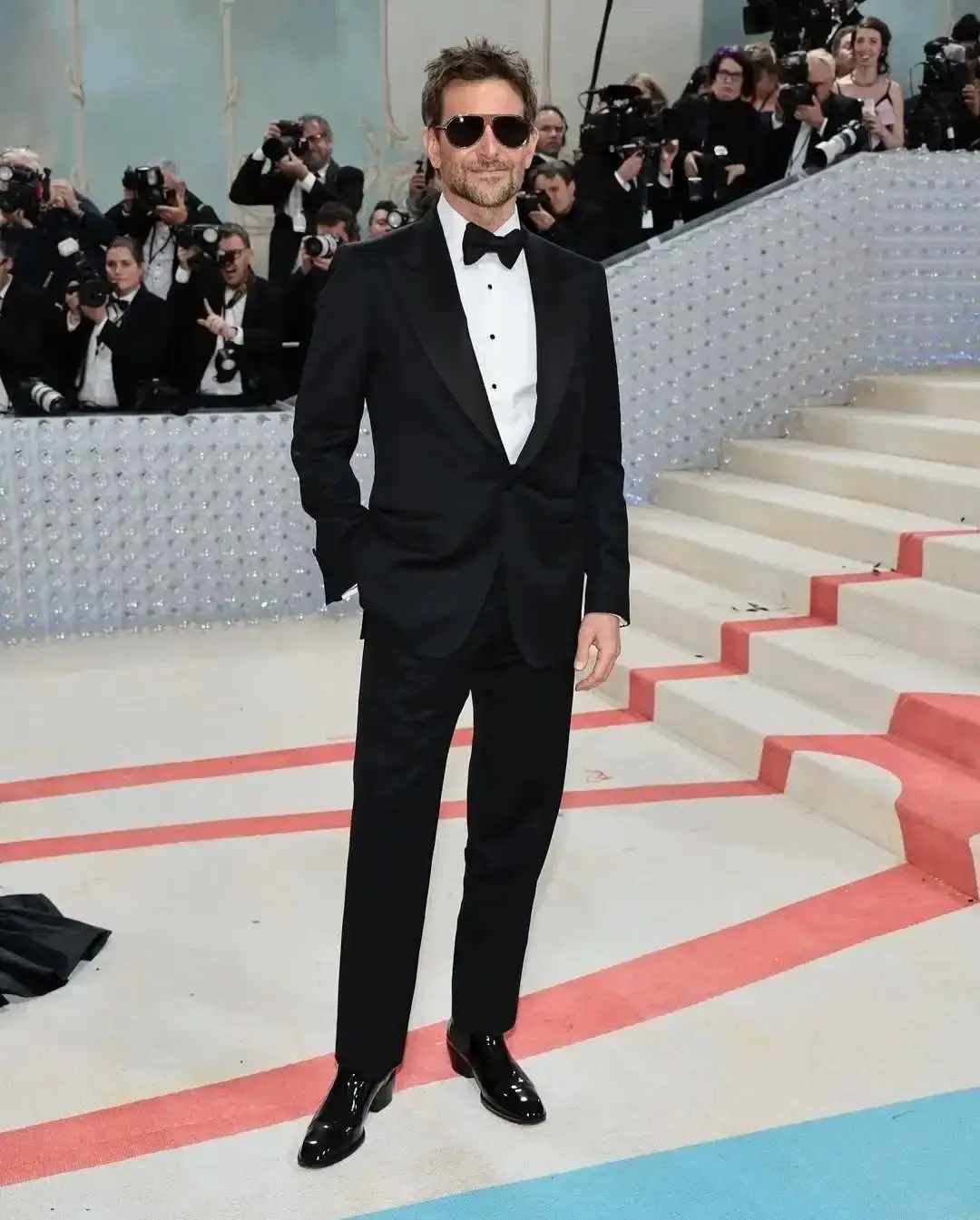 Actor Bradley Cooper in a black tuxedo and bow tie with sunglasses, posing formally on a red carpet with a staircase and a crowd of photographers behind him.