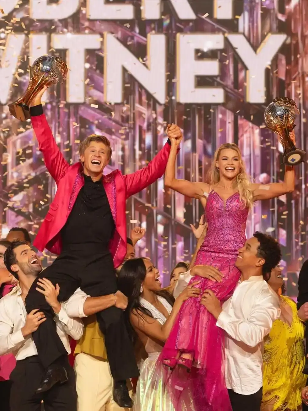 Robert Irwin and Witney Carson are lifted onto the shoulders of other dancers, celebrating a victory and holding up mirrorball trophies amidst falling confetti.
