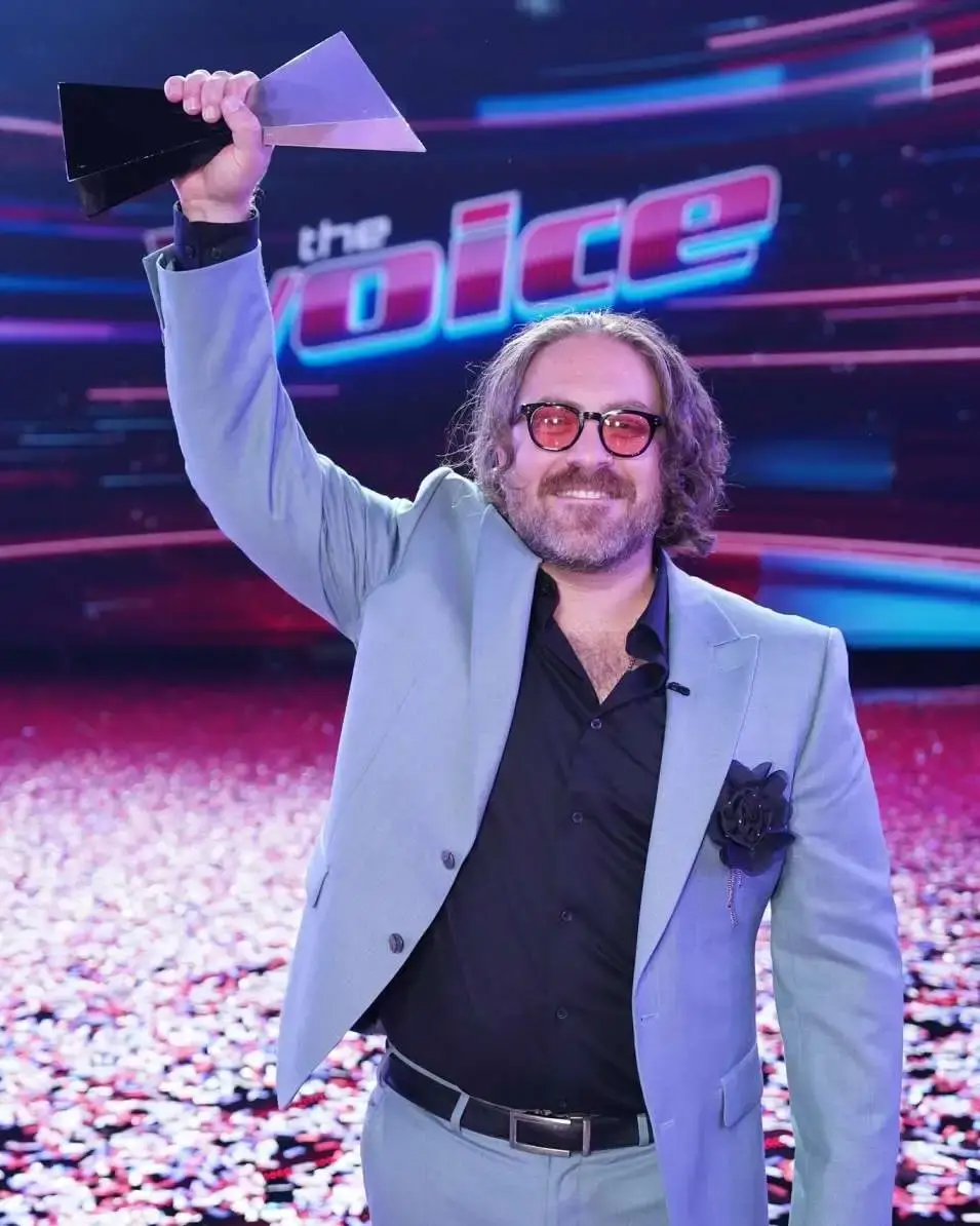 Singer Adam David, wearing a light blue suit and red sunglasses, raises a triangular trophy on stage with "The Voice" logo visible in the background.