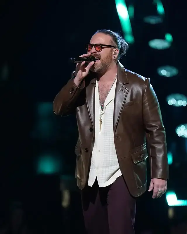 Singer Adam David, with his hair tied back, performs on stage in a brown leather blazer, a white collared shirt, and sunglasses, holding a microphone to his mouth.