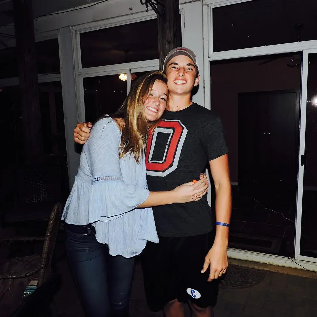 College quarterback Max Brosmer in a dark Ohio Buckeyes t-shirt is hugged by his cousin, Libby Wehr, outside at night, with both smiling for the camera.