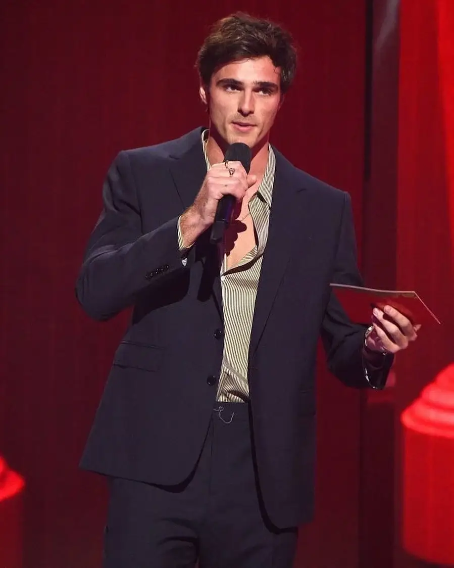 Actor Jacob Elordi on a stage in a dark navy suit and an unbuttoned tan striped shirt, holding a microphone and a red cue card.