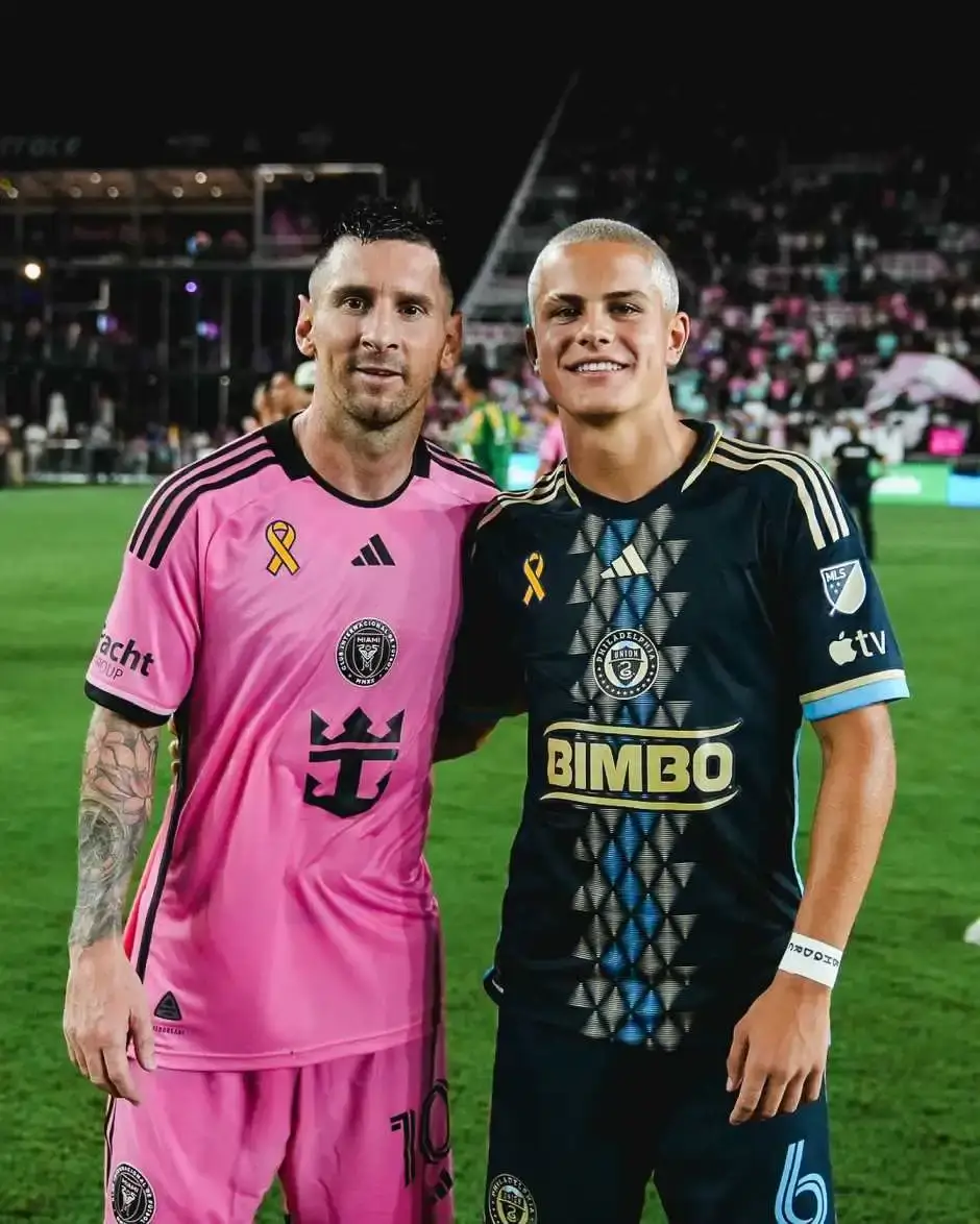 Soccer star Lionel Messi in a pink Inter Miami jersey smiles next to young player Cavan Sullivan in a dark Philadelphia Union jersey on a pitch.