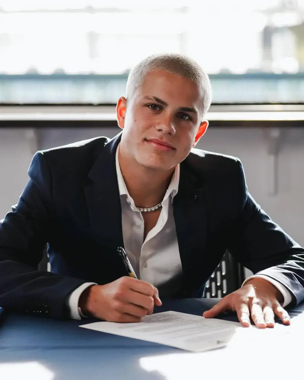 Young soccer player Cavan Sullivan, with a shaved blonde head, sits at a table signing a contract while wearing a dark suit jacket and a diamond necklace.