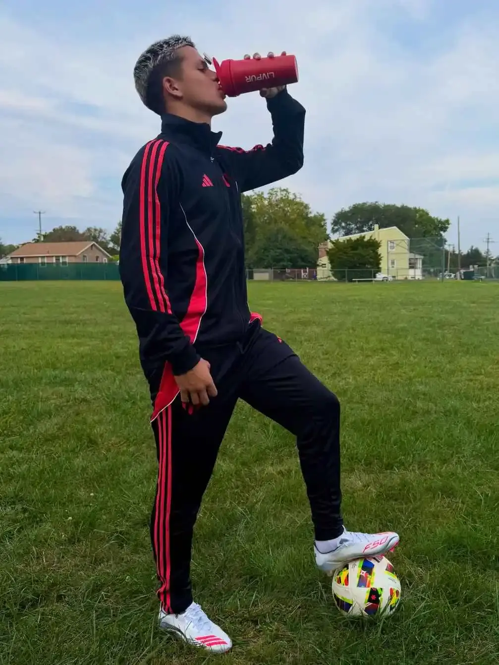 Young soccer player Cavan Sullivan, wearing a black and red Adidas tracksuit, drinks from a red water bottle while stepping on a soccer ball outdoors.