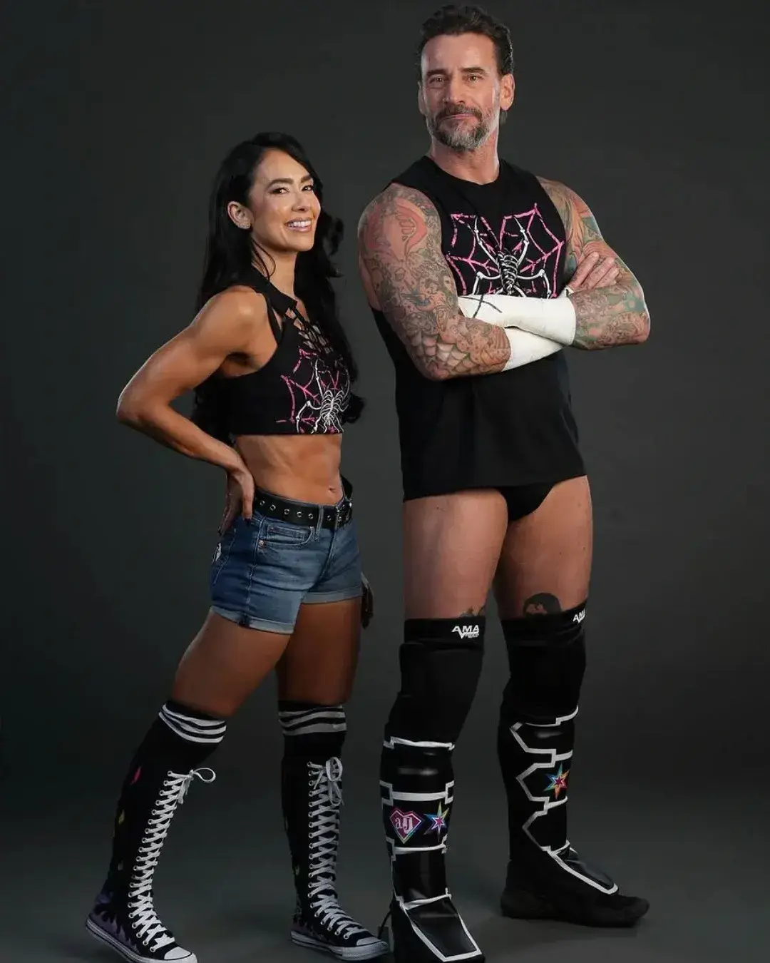 : Professional wrestlers CM Punk with crossed arms and his wife AJ Lee, who is wearing cutoff denim shorts and a spiderweb top, pose together on a gray background.