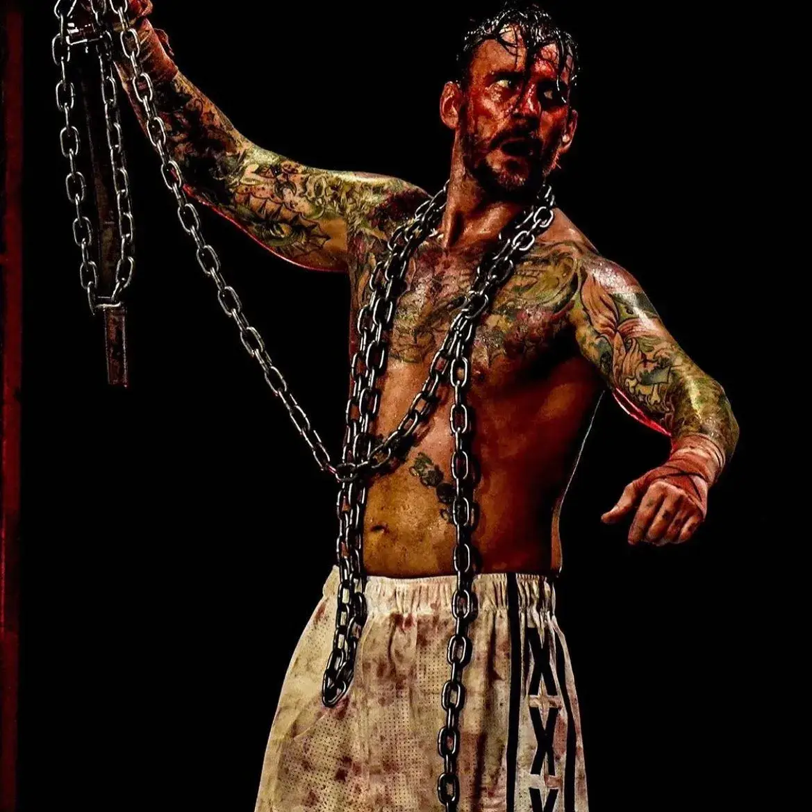 Professional wrestler CM Punk, covered in blood with chains draped over his tattooed torso, holds a chain up against a dark background.
