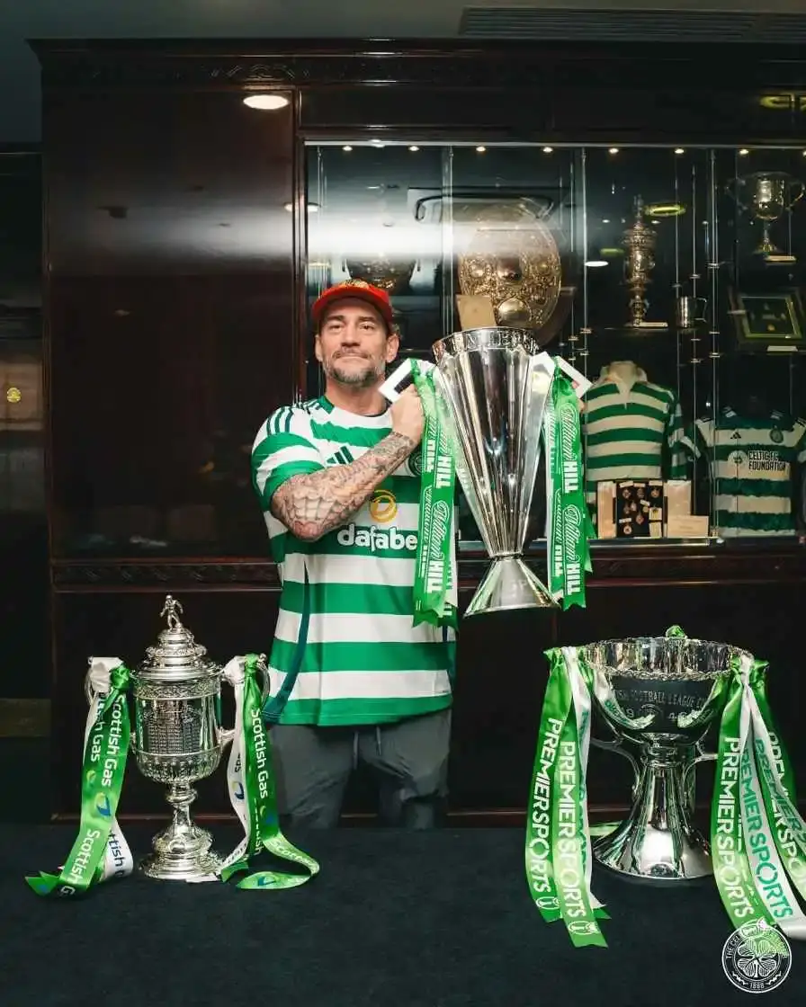 Professional wrestler CM Punk in a Celtic FC jersey and a red baseball cap stands in front of a trophy case, holding a large silver trophy.