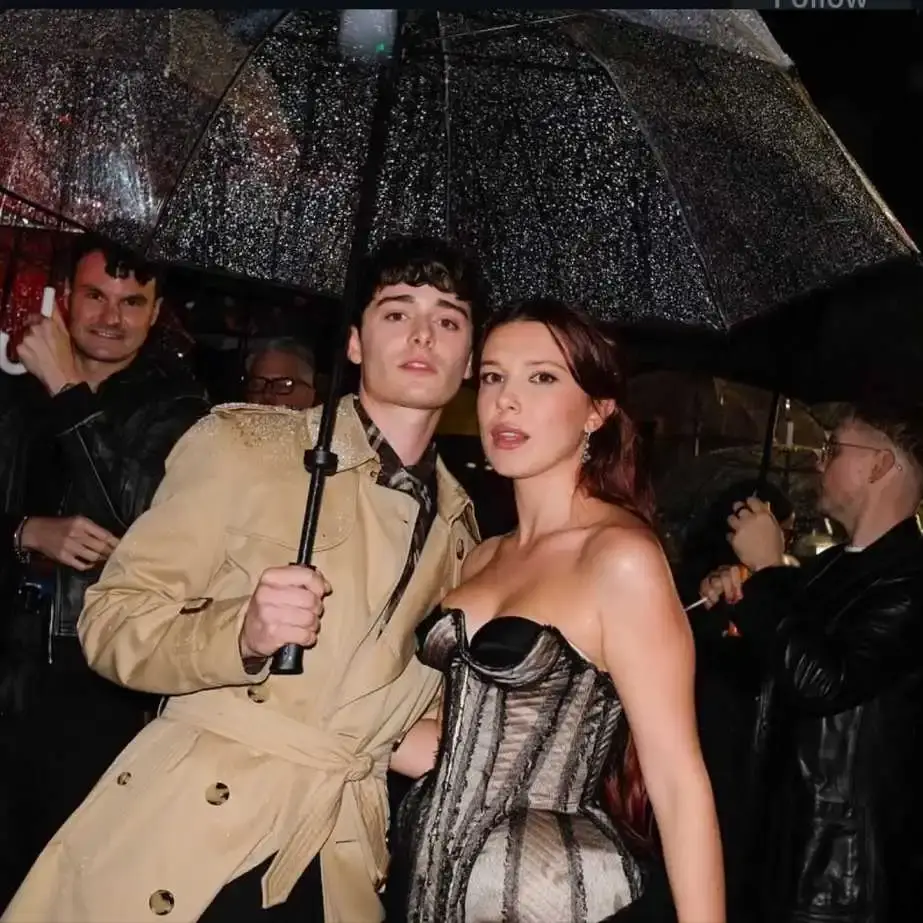 Actors Noah Schnapp and Millie Bobby Brown standing close together under a clear umbrella in the rain, with Millie Bobby Brown wearing a strapless corset and Noah Schnapp wearing a trench coat.