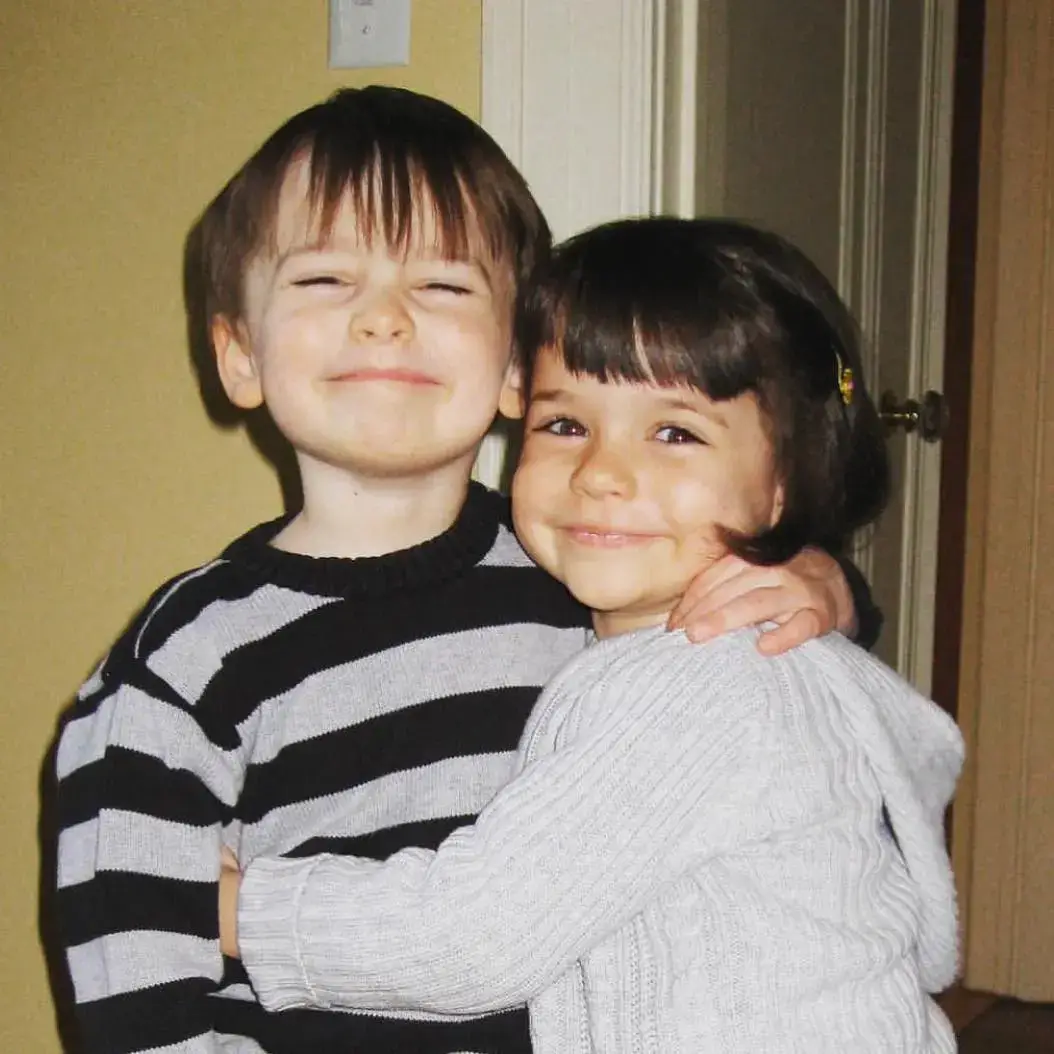 A childhood photo of twin siblings Noah Schnapp in a striped sweater and Chloe Schnapp in a grey sweater, hugging and smiling sweetly at the camera.