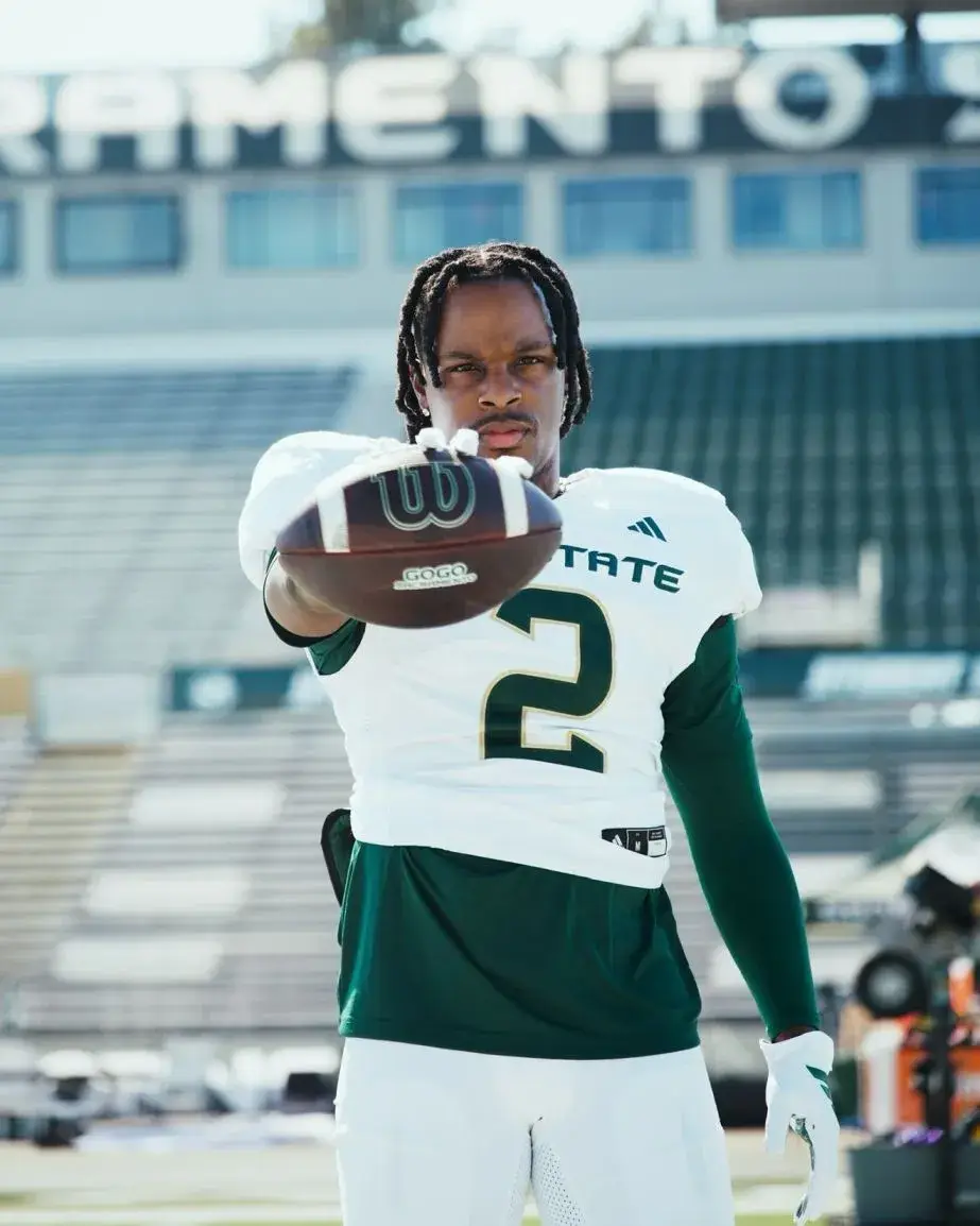 Musician Toosii, with dreadlocks, posing on a football field in a white and green football uniform, holding a football directly towards the camera.