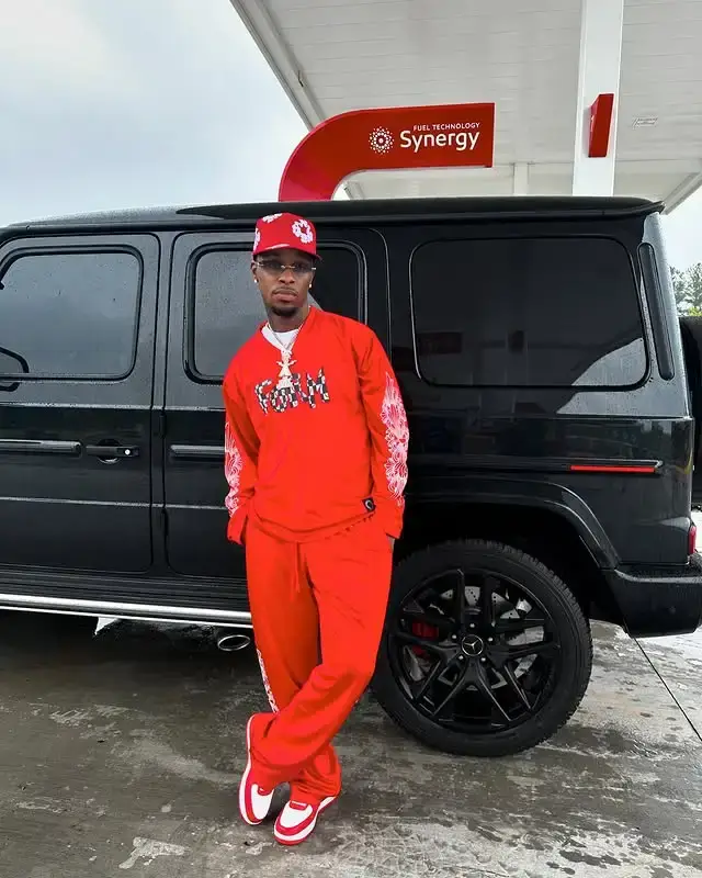 Musician Toosii posing in a full red tracksuit, a red cap, and sneakers, leaning on a black Mercedes-Benz G-Wagen at a gas station.