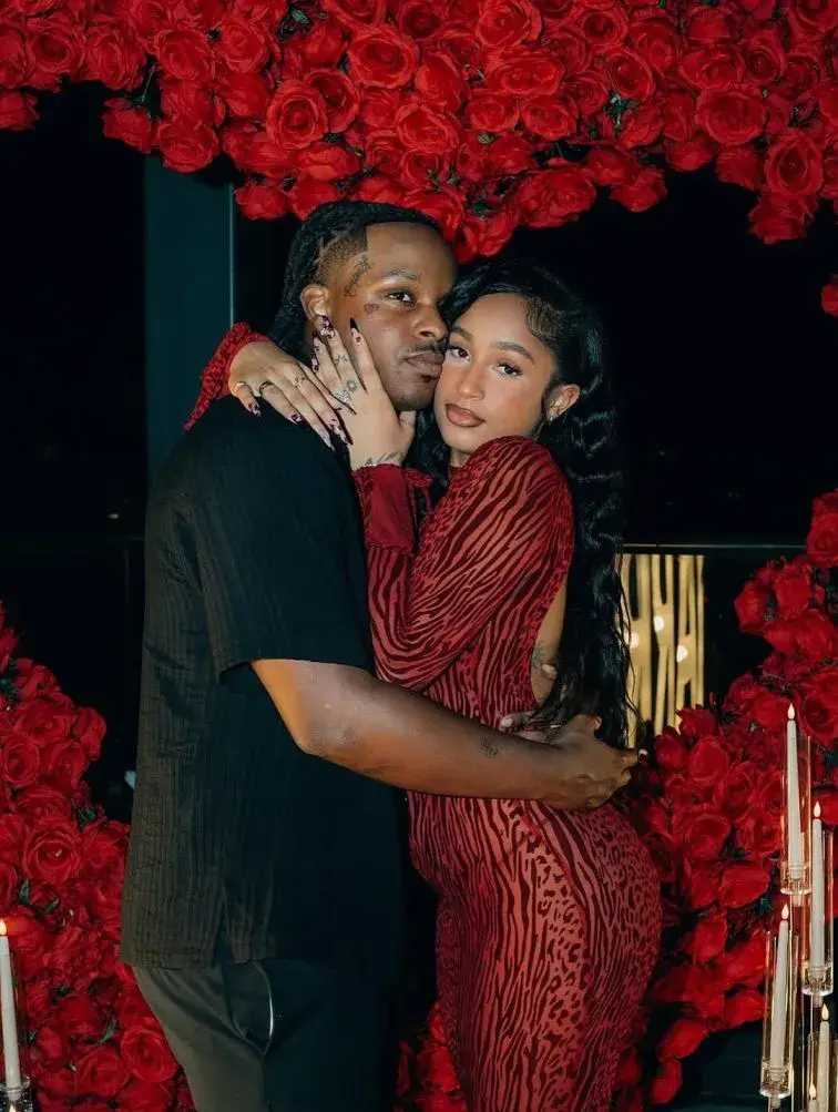 Rapper Toosii and his girlfriend Samaria J. Davis are embracing in front of a wall of red roses and candles on a dark background.