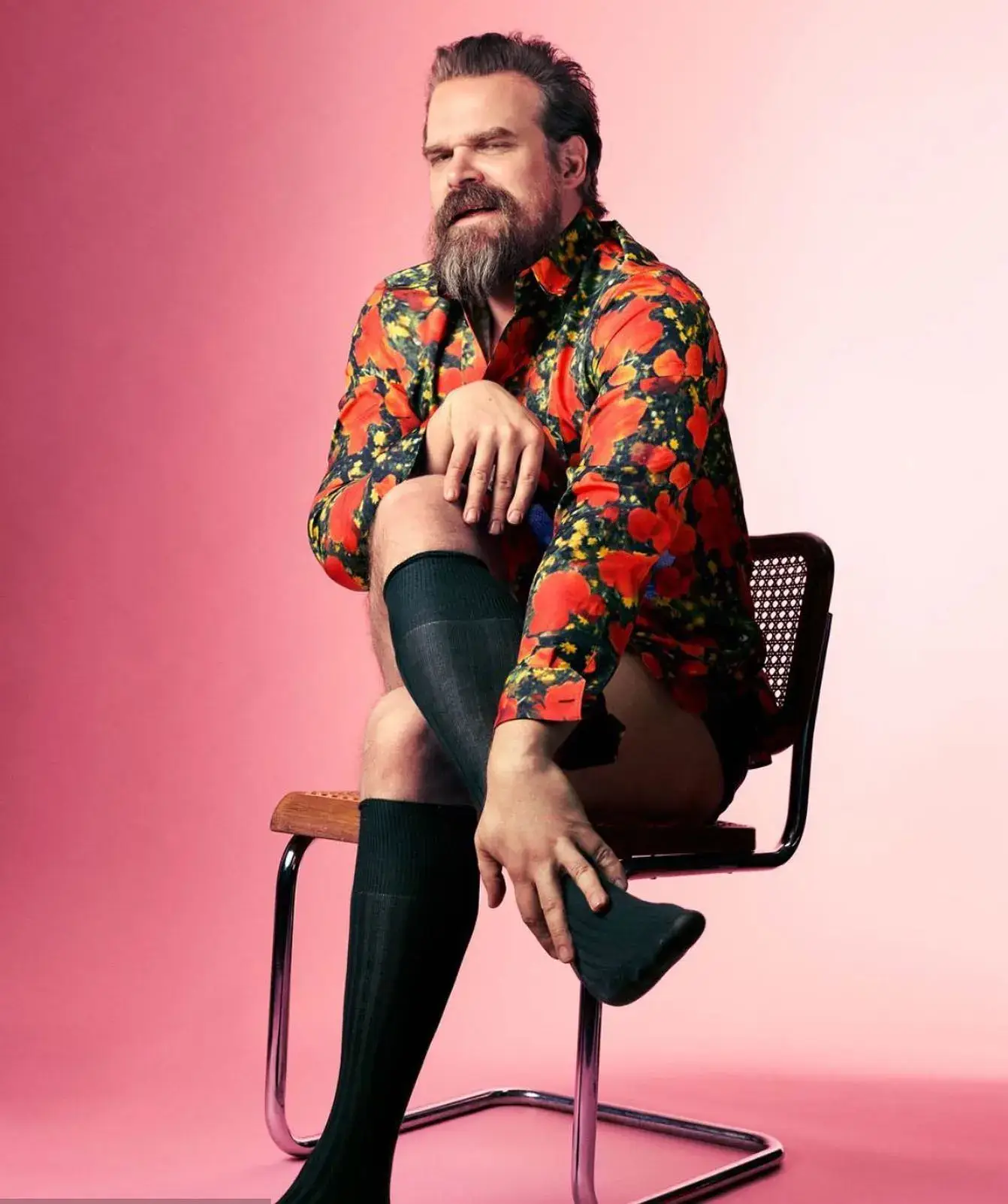 Actor David Harbour in a bright floral print shirt and black socks pulled up to his knees, sitting on a chair against a pink background.