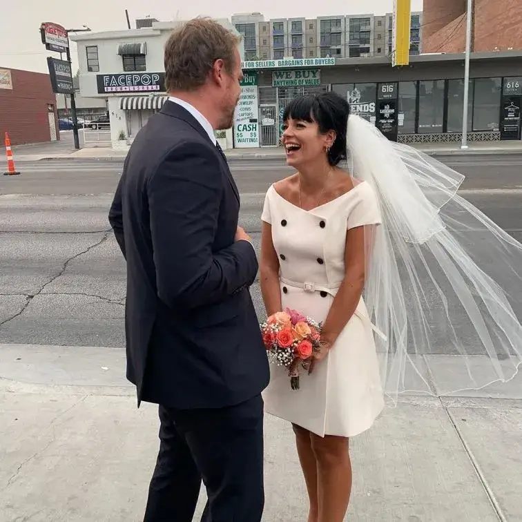 David Harbour in a black tuxedo and Lily Allen in a short white wedding dress are smiling next to an Elvis impersonator in a gold jumpsuit in Las Vegas.