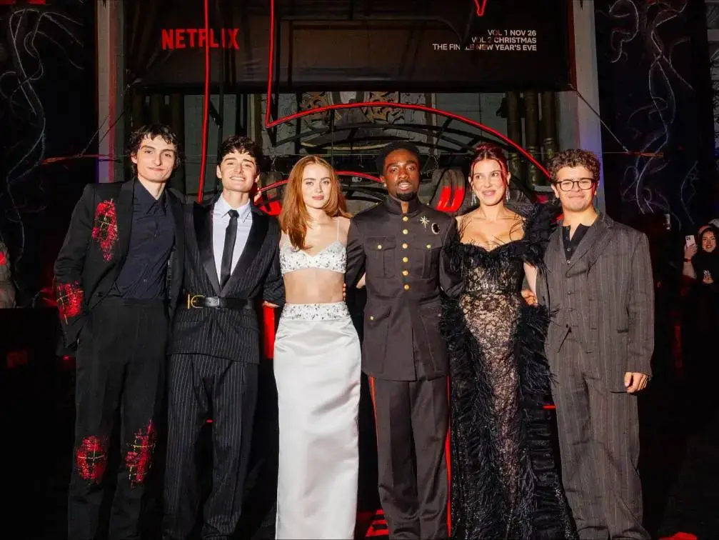 A group photo of five Stranger Things actors, including Finn Wolfhard, Noah Schnapp, Sadie Sink, Caleb McLaughlin, and Natalia Dyer, posing formally on a red carpet.