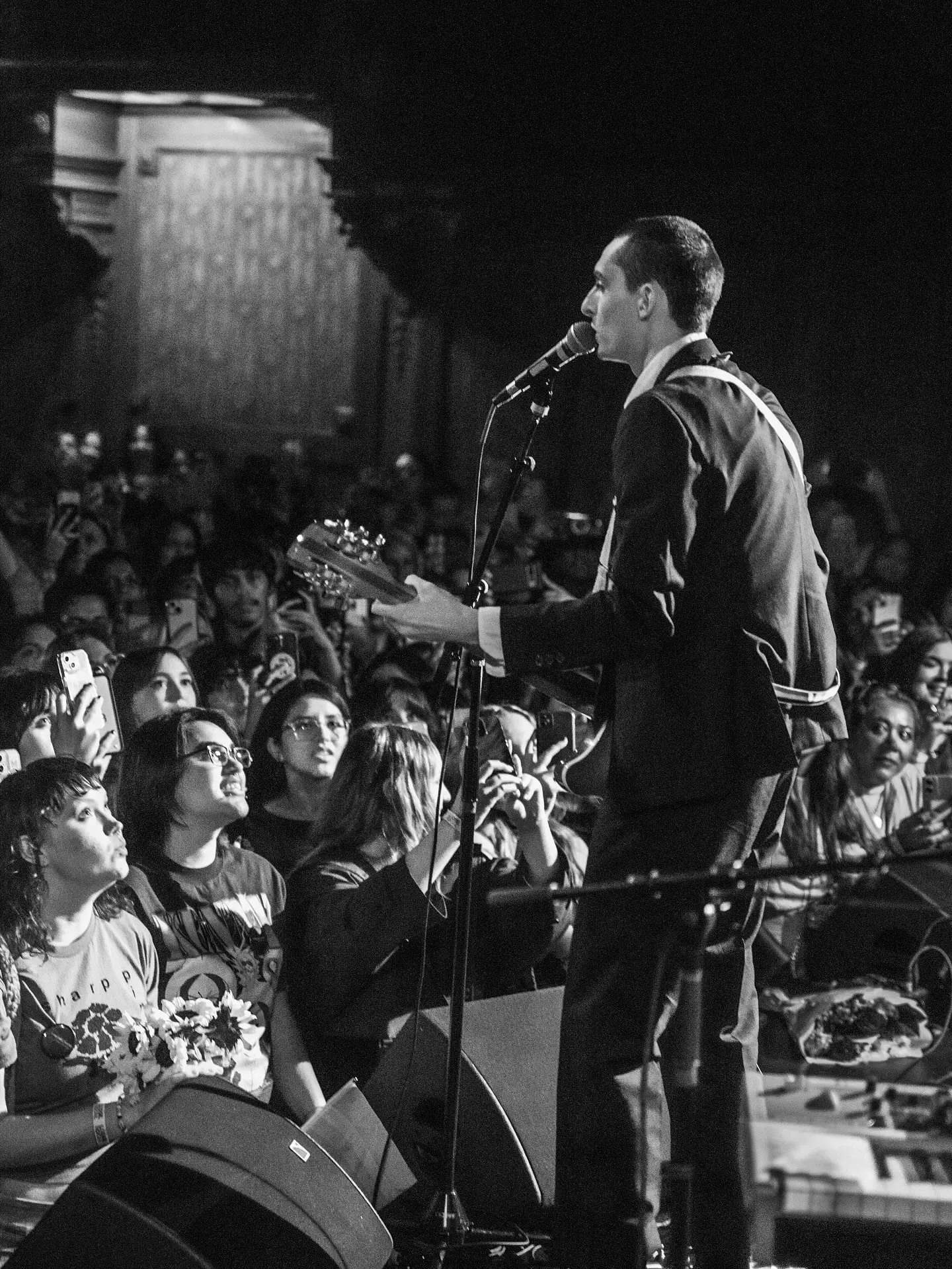 Black and white full body shot of actor Finn Wolfhard performing live music on stage with an electric guitar in a dark suit to a crowd of cheering fans.