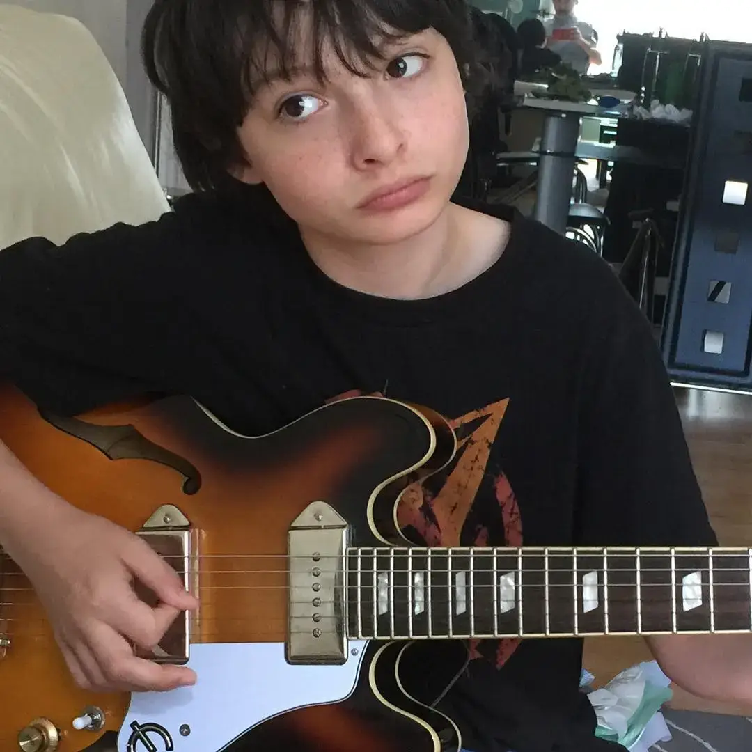 Young Finn Wolfhard with a thoughtful expression looking off to the side while holding an electric guitar in a close-up picture.