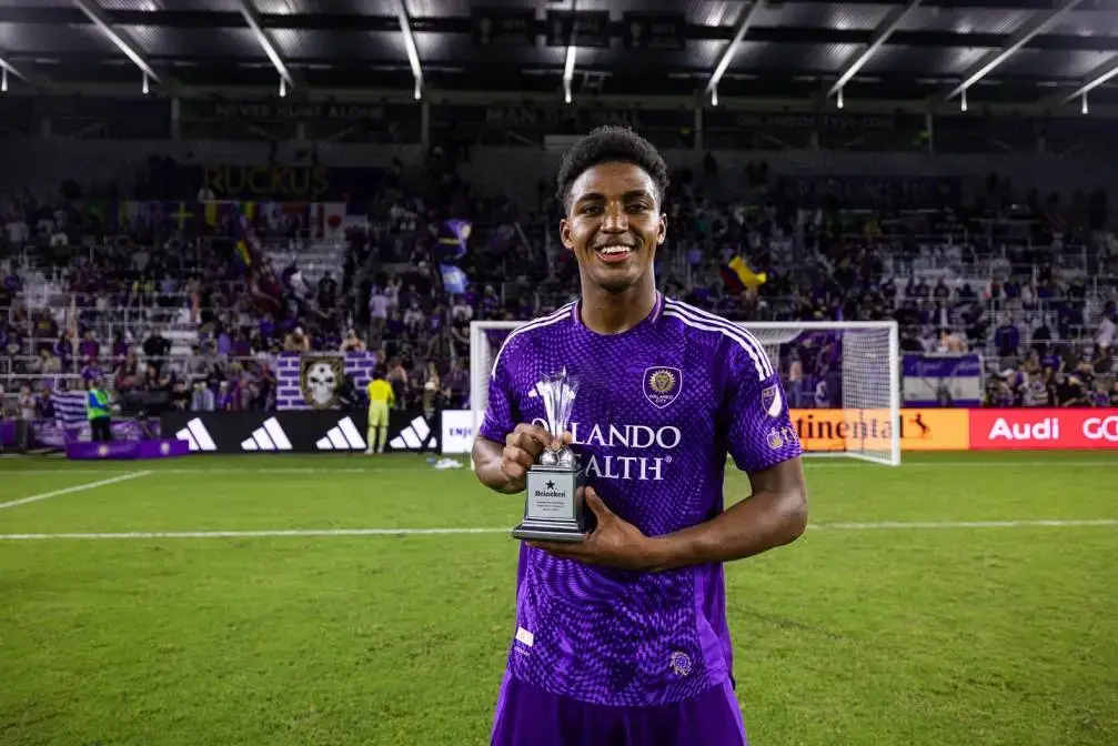Soccer player Alex Freeman in a purple Orlando City SC jersey stands on the field at night holding a small silver trophy with a celebratory smile.