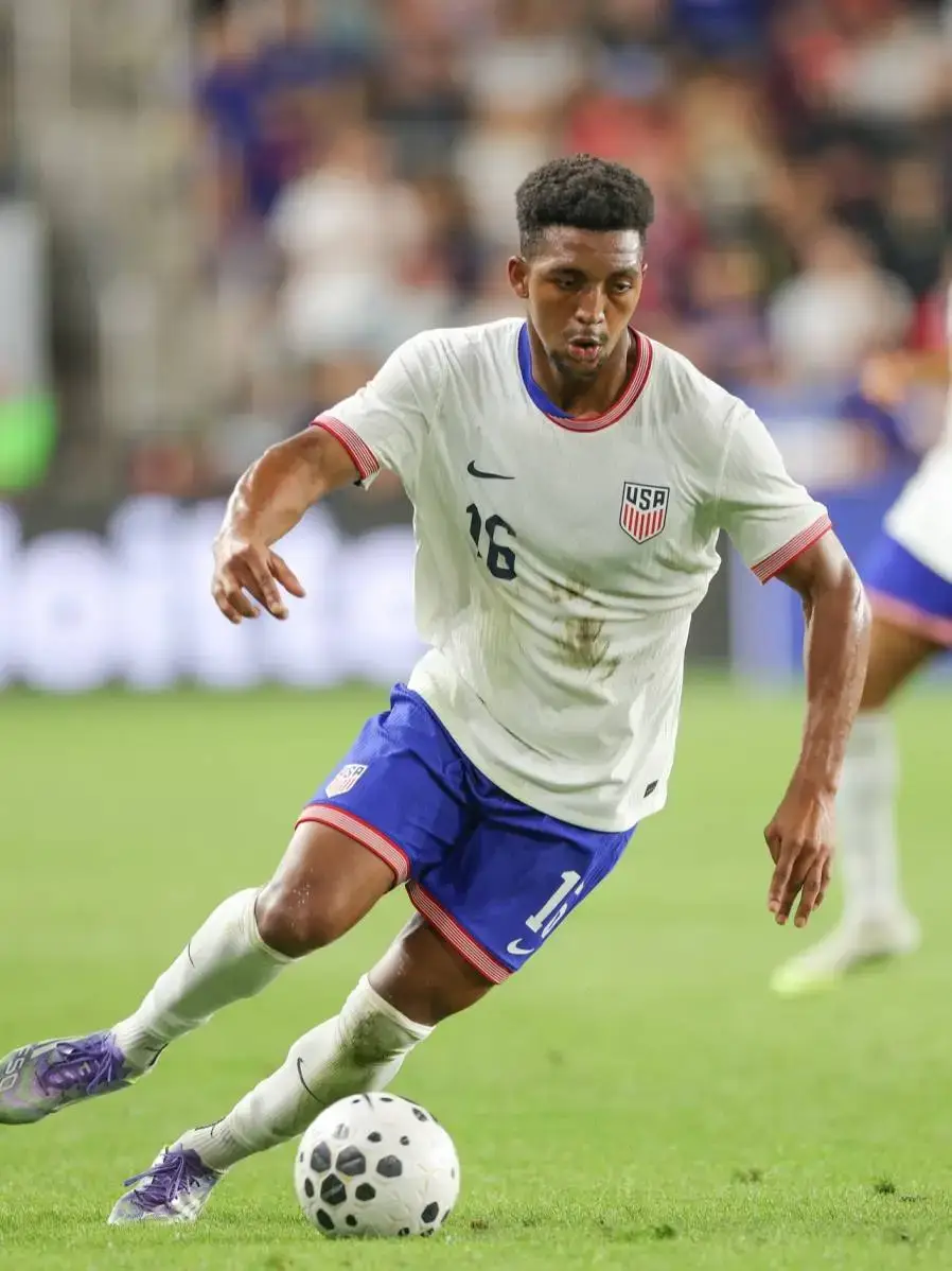 Soccer player Alex Freeman in a white US National Team jersey with the number 16 on the field, dribbling a black and white patterned soccer ball.