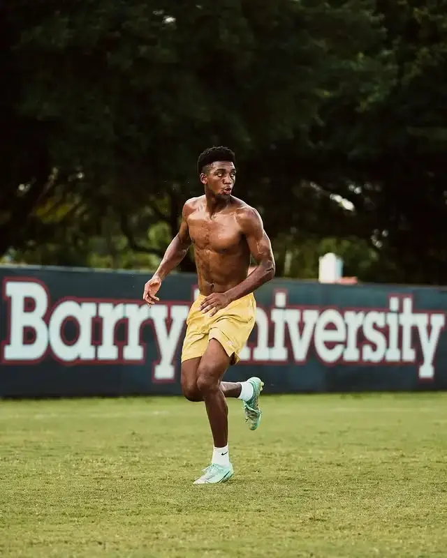 Shirtless soccer player Alex Freeman runs across a green field in yellow shorts and light-colored cleats during a training session with "Barry University" visible behind him.