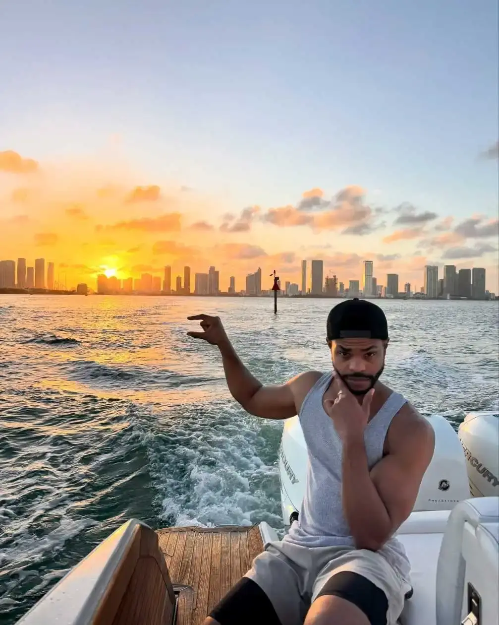 Comedian and actor King Bach sits on the back of a boat at sunset with the Miami skyline in the background, pointing toward the city.