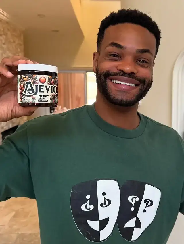 Comedian and actor King Bach smiling while holding a jar of supplements, wearing a dark green t-shirt with two drama masks on it.