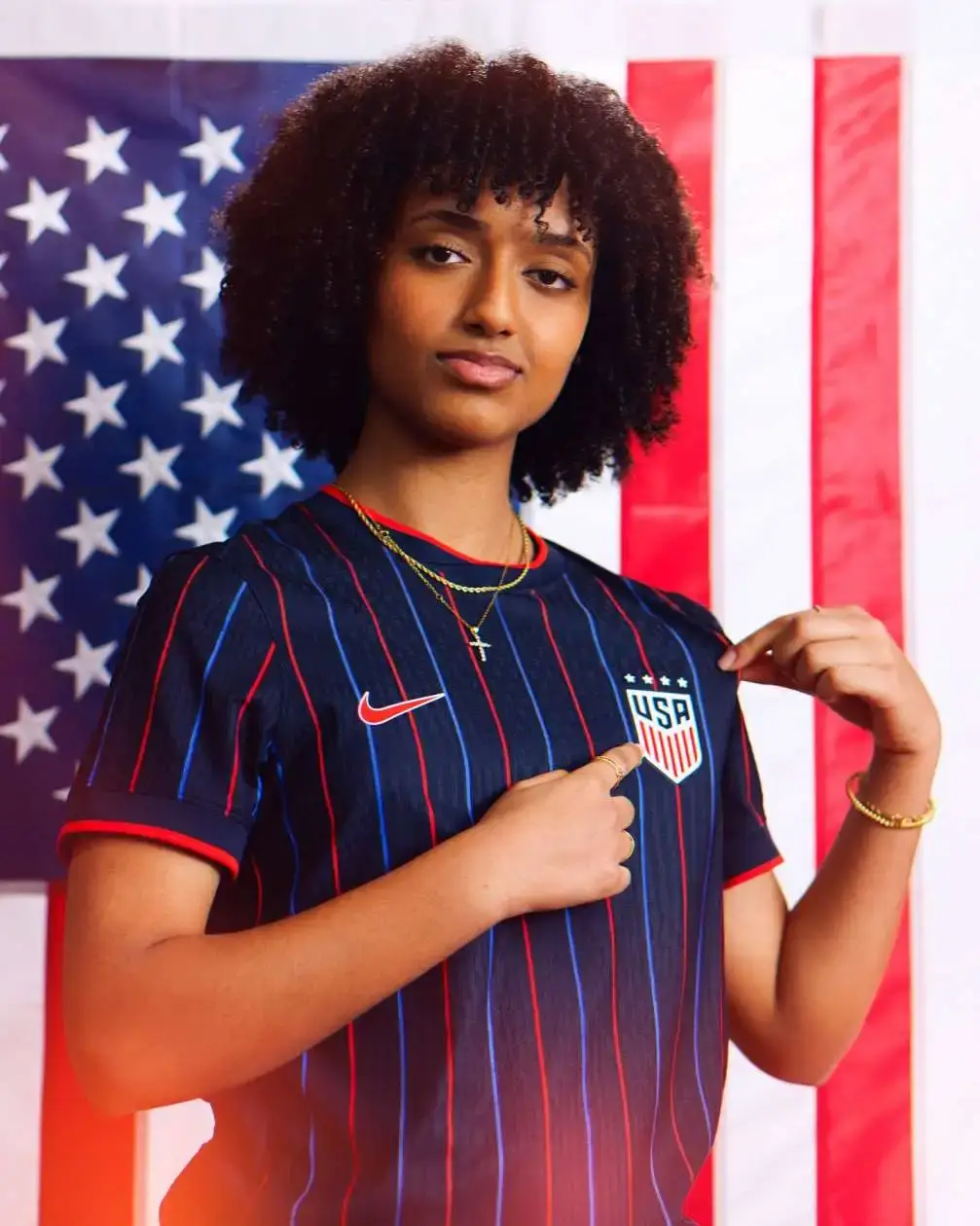 Soccer player Lily Yohannes stands in front of an American flag, pointing to the USA crest on her blue striped national team jersey.