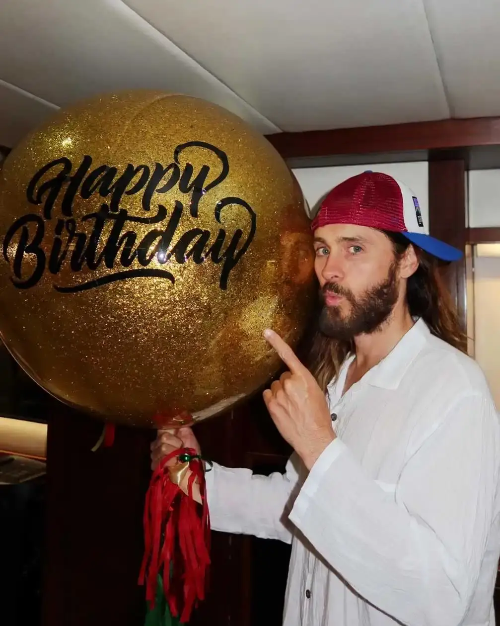 Actor and musician Jared Leto wearing a white shirt and a baseball cap backward, pointing to a large, round, gold "Happy Birthday" balloon.