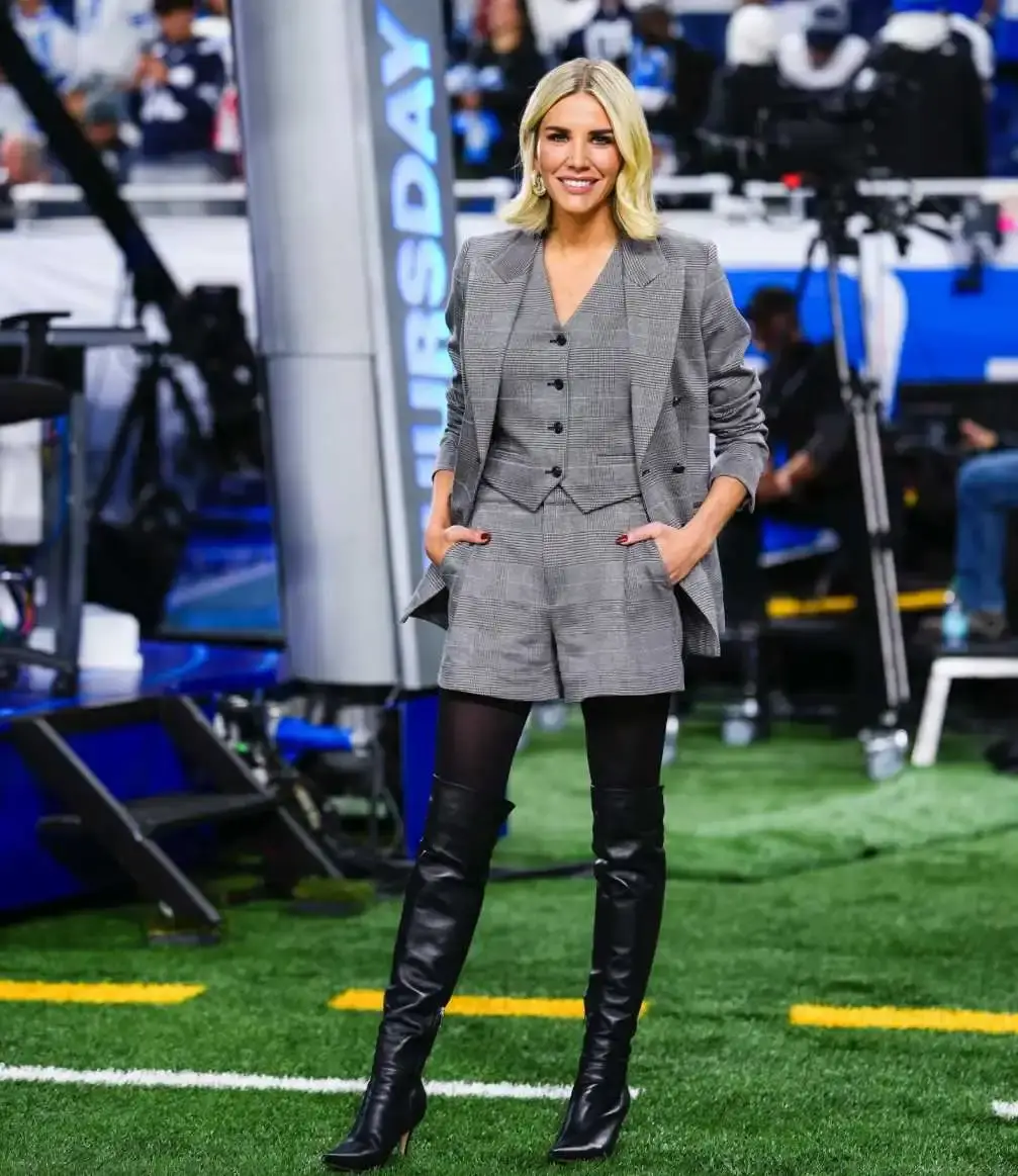 Sports reporter Charissa Thompson posing on a football field in a gray plaid shorts suit with a vest, a blazer, tights, and black thigh-high boots.