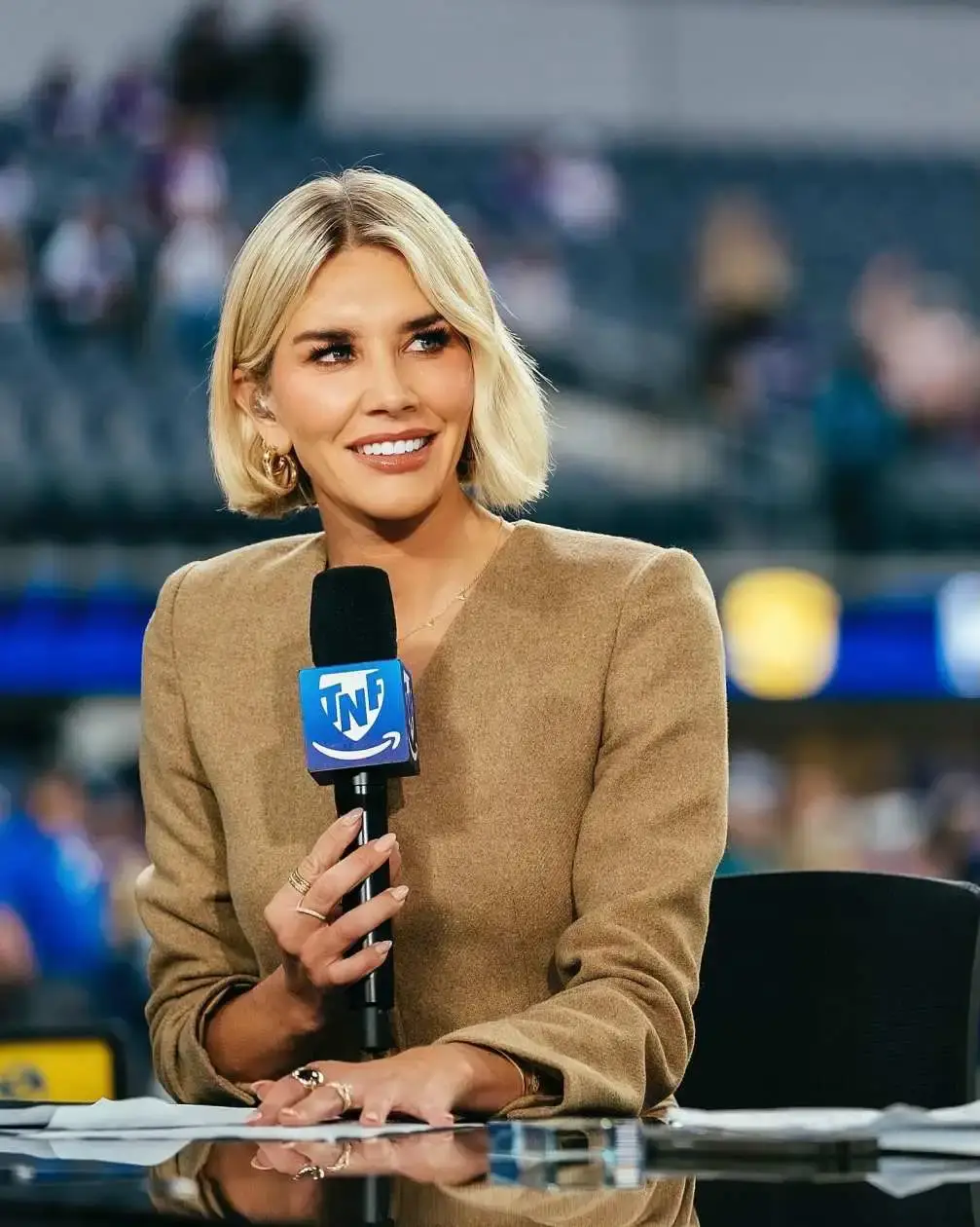 Sports reporter Charissa Thompson with blonde hair and a beige blazer smiles while holding a TNT Sports microphone at a desk on the sidelines of a game.