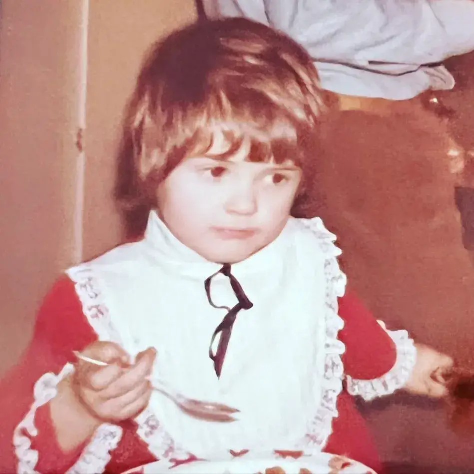 A childhood photo of Dagen McDowell with bangs, wearing a red top and a white bib with lace trim, holding a spoon while looking to the side.