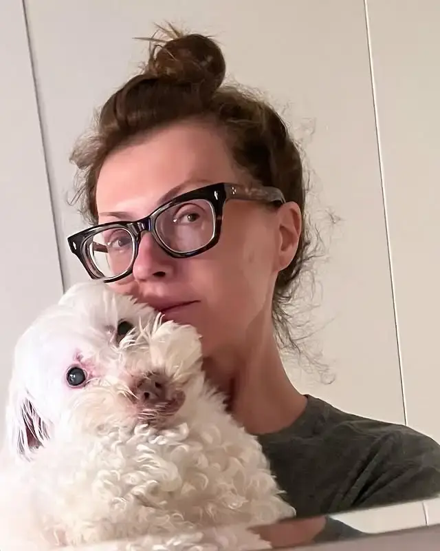 Dagen McDowell, wearing glasses, holds a small white fluffy dog named Ramon close for a mirror selfie indoors.