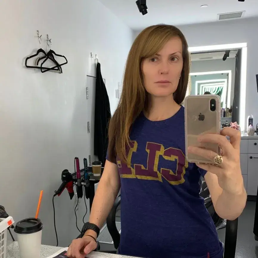 Financial journalist Dagen McDowell, with long reddish-brown hair, takes a mirror selfie in a casual blue t-shirt in a hair and makeup room.