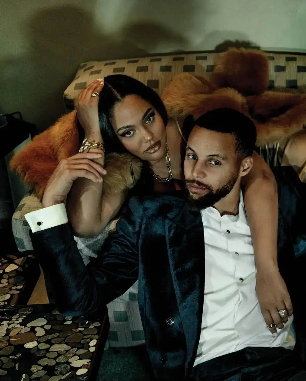 Steph Curry and his wife Ayesha Curry pose closely on a couch, with Ayesha wearing a fur stole and jewelry and resting her arm over Steph