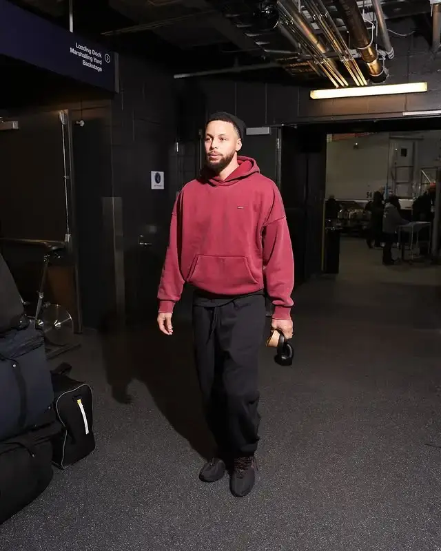 NBA superstar Steph Curry walks in a dark corridor wearing a maroon hoodie and black sweatpants.