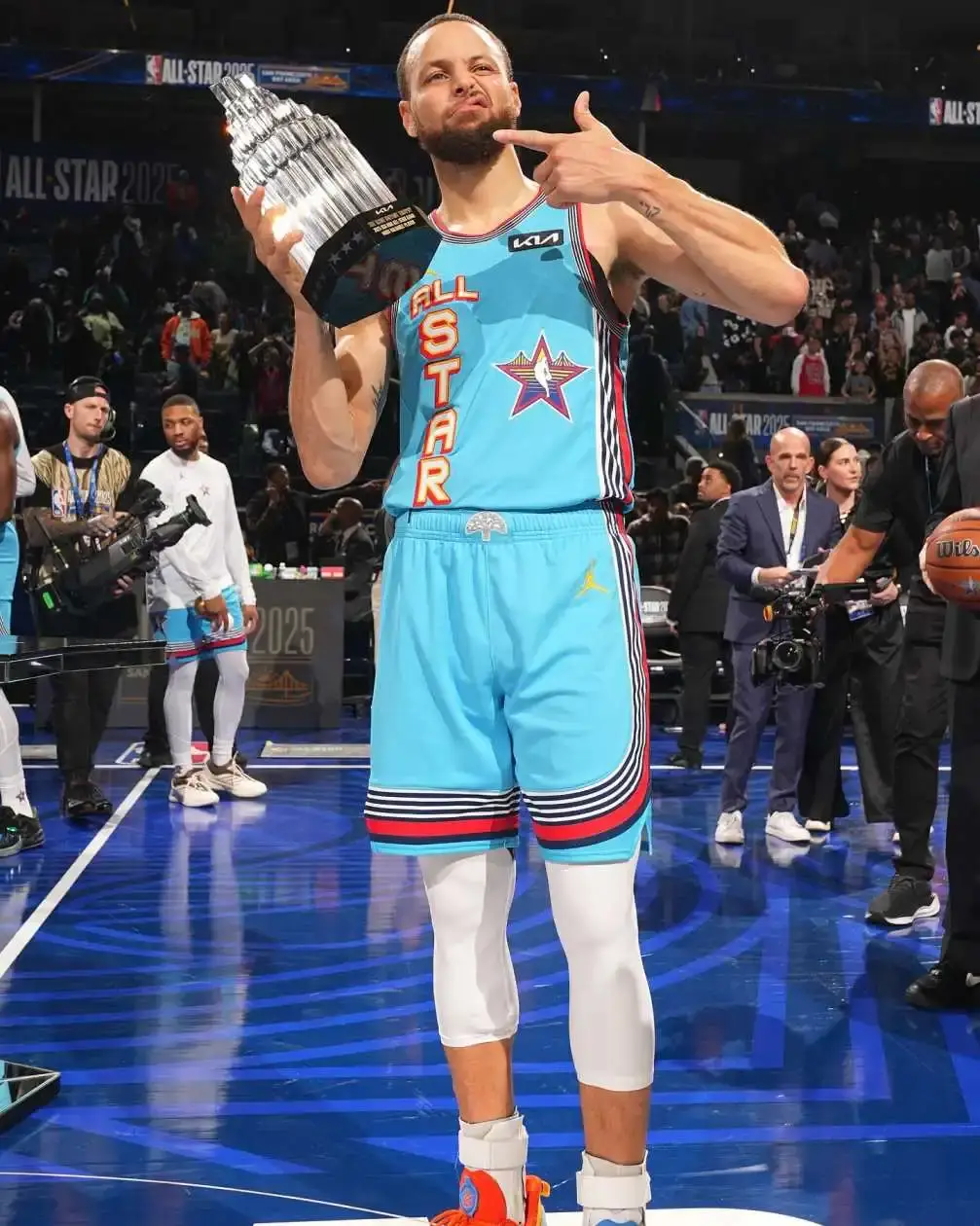 NBA player Steph Curry holds the All-Star MVP trophy high while standing on the court in a blue All-Star uniform and pointing at the trophy.