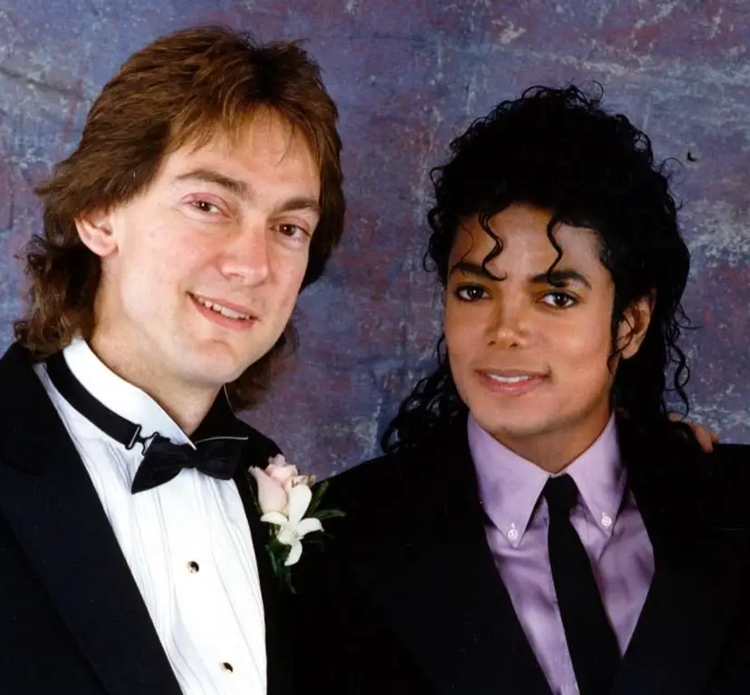 John Branca in a tuxedo next to Michael Jackson in a black suit with a lavender shirt and black tie, posing for a portrait against a mottled background.