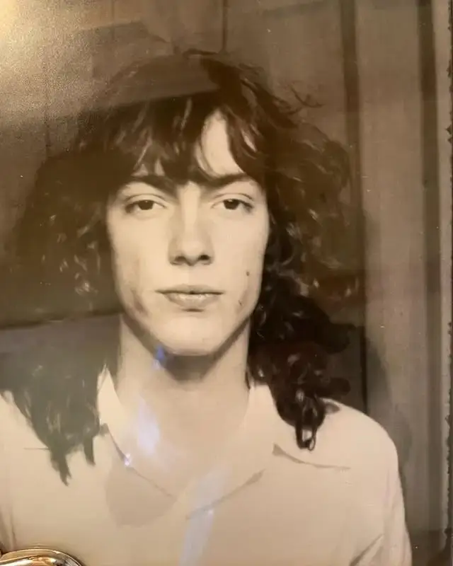 A black and white portrait of a young John Branca at age 16 with long, curly dark hair and a serious expression, wearing a white collared shirt.