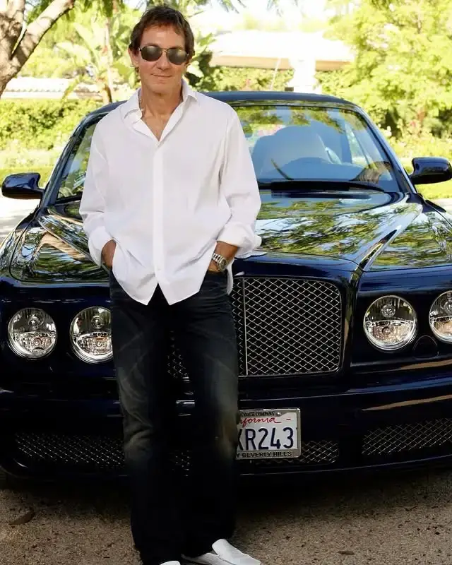 Music attorney John Branca, dressed casually in a white button-down shirt and dark jeans, stands in front of a shiny dark blue Bentley convertible.