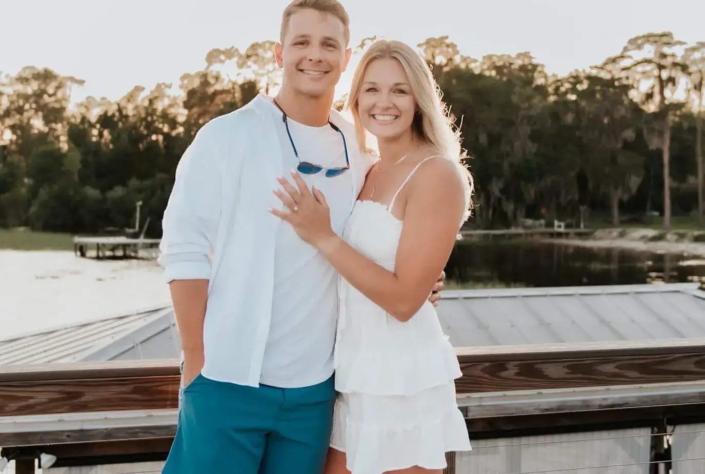 NFL player Brock Purdy and his wife Jenna Purdy smiling for a photo, with Jenna showing off an engagement ring on her left hand in front of a lake backdrop.