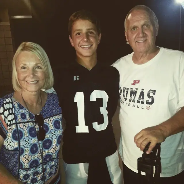 Young Brock Purdy in a black football jersey stands between his smiling blonde-haired grandmother and his grandfather in a white "Pumas Football" shirt.