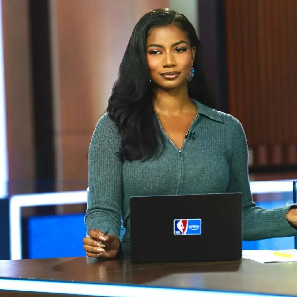 Sports journalist Taylor Rooks with long dark hair, wearing a gray zippered collared top, sits at a desk with a laptop bearing the NBA and Amazon Prime logos.