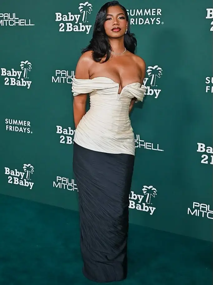 Sports journalist Taylor Rooks stands on a red carpet wearing a white off-the-shoulder corset top and a long, flowing black skirt, posing in front of a green backdrop.