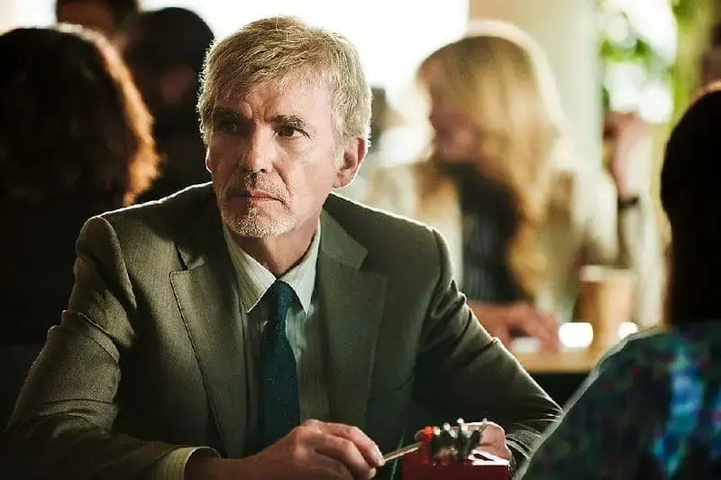 Actor Billy Bob Thornton looks intently at someone off-camera while sitting at a table in a suit jacket and tie for a film role.