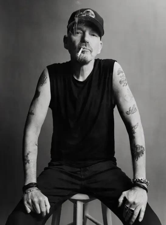 Black and white portrait of actor Billy Bob Thornton sitting on a stool, wearing a sleeveless shirt, a baseball cap, showing his arm tattoos, and holding a cigarette in his mouth.