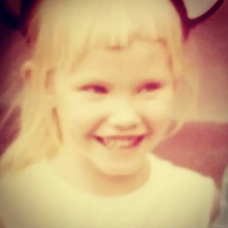 A slightly faded, soft-focus photo of a young, blonde-haired Kelli Giddish with bangs, smiling broadly at the camera.